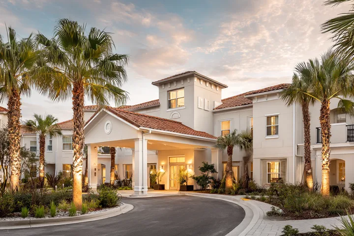 A two-story cream stucco senior living residence with a covered entry and a circular drive among palms at dusk