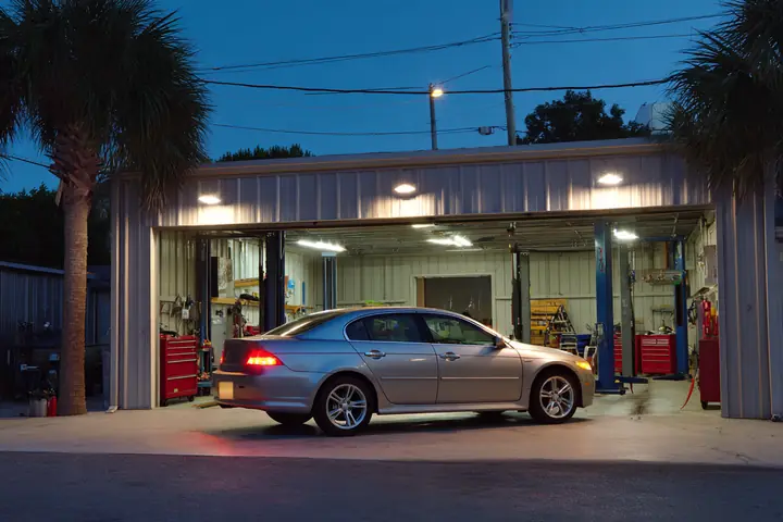An open service bay at dusk, a sedan raised on a two-post lift under bright shop lights, tools along the back wall