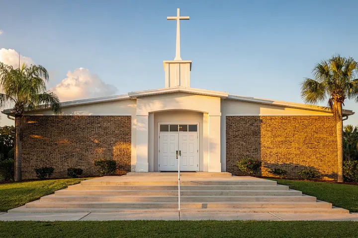 A brick and white church with a tall white steeple and cross, wide front steps and double doors between palms in late afternoon light