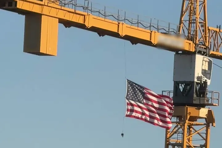 A yellow tower crane flying an American flag against a clear blue sky over a construction site in downtown Naples, Florida