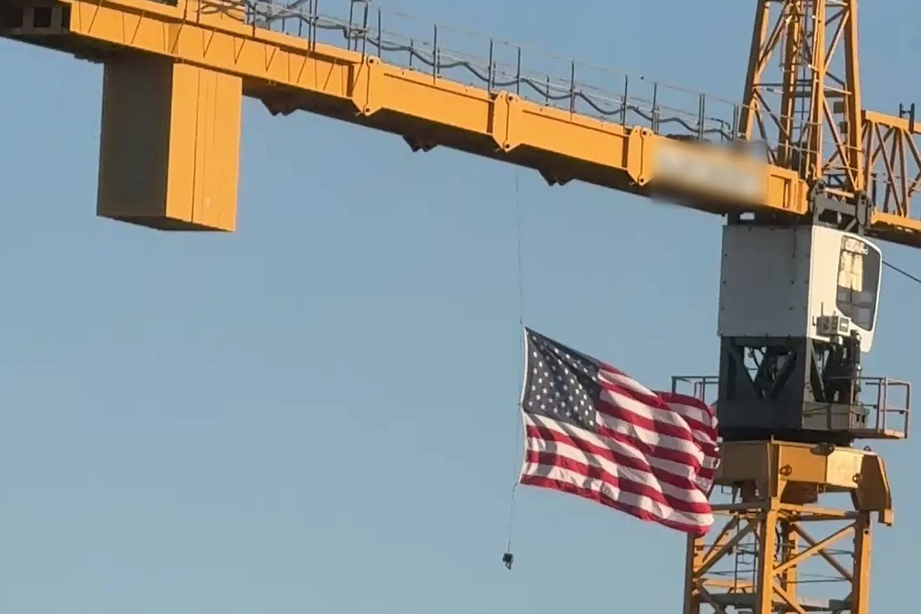 A yellow tower crane flying an American flag against a clear blue sky over a construction site in downtown Naples, Florida