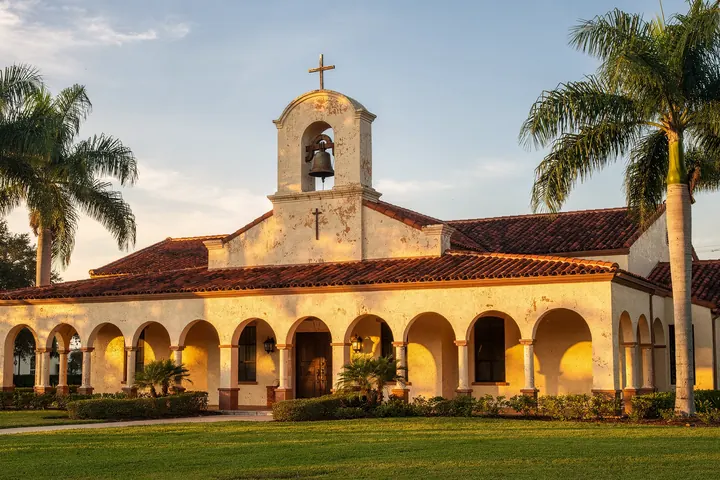 A cream stucco mission style church with a bell tower, a plain cross and an arched loggia under royal palms in late afternoon light