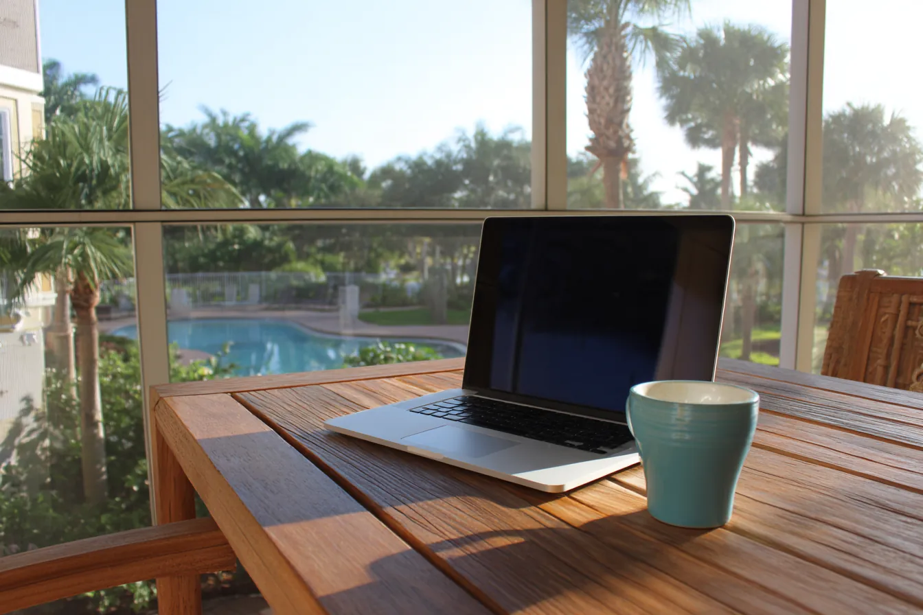 A closed laptop on a teak table on a screened Florida lanai, a pool and palms beyond in morning light
