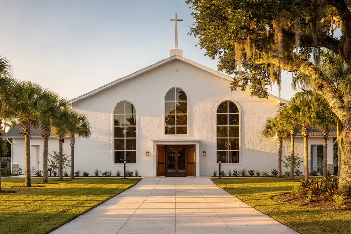 A white stucco church with a plain cross on its steeple, arched windows and open wooden doors, palms and a live oak in late afternoon light