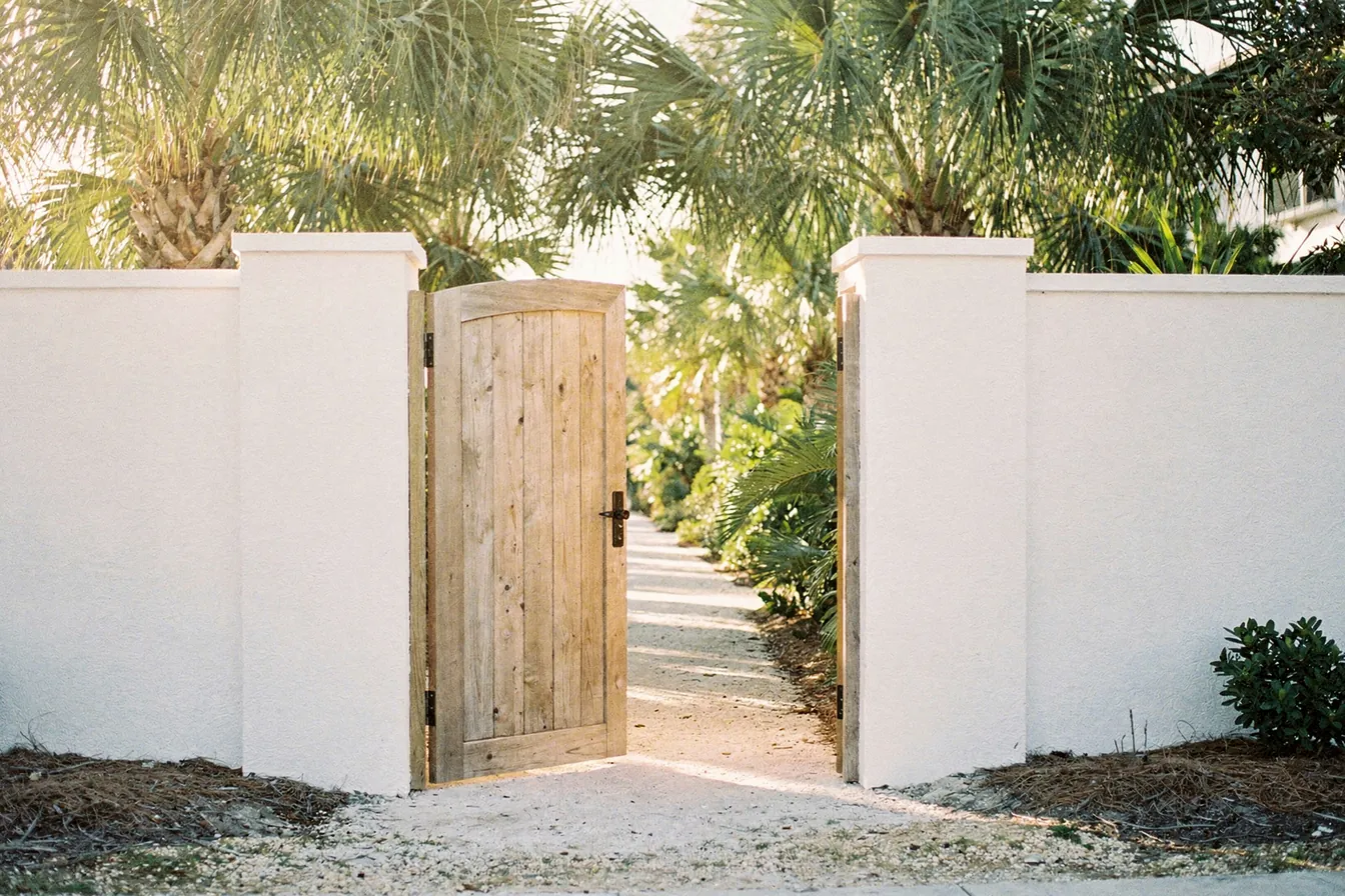 An open wooden gate in a white stucco garden wall leading to a sunny path