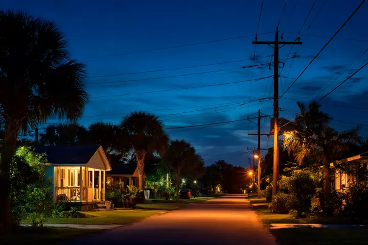 A Southwest Florida street at blue hour, porch lights and windows glowing, palms and a utility pole silhouetted against the sky