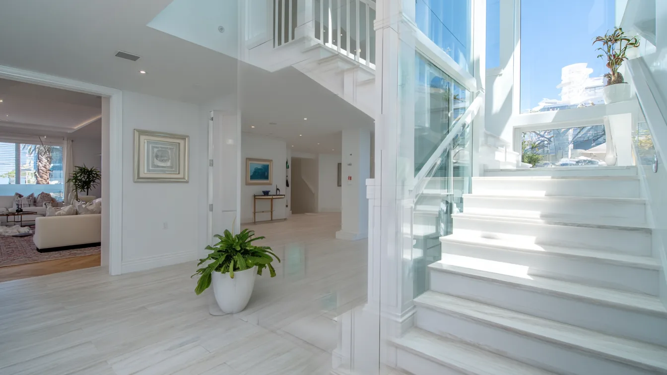 A bright two-story Southwest Florida foyer seen straight on: a doorway to the living room on the left, a plain white wall in the middle and a white staircase with a glass railing on the right