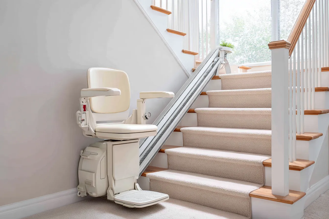 A stair lift chair folded at the foot of a rail running up a carpeted staircase in a bright, white home