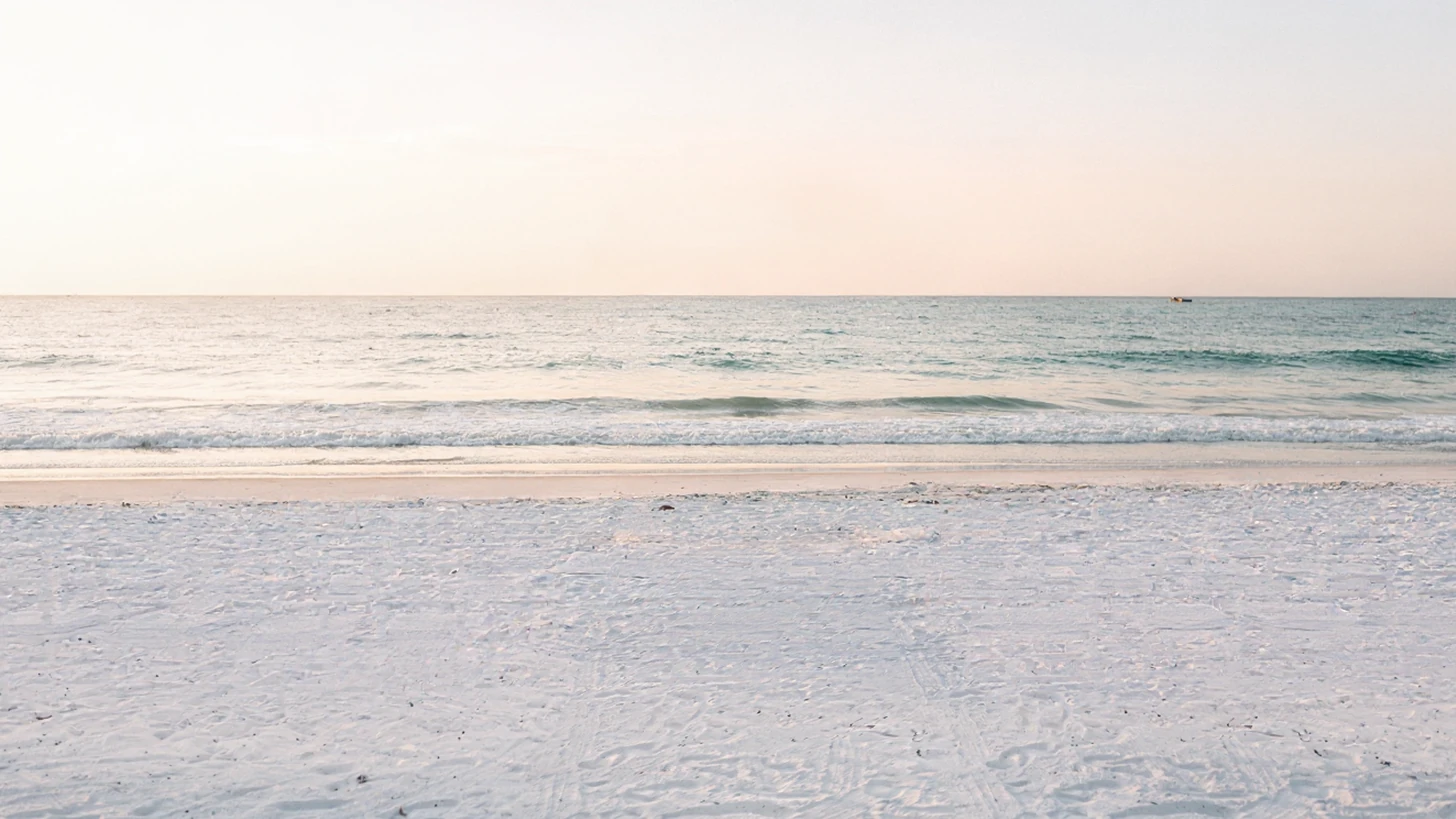 An empty stretch of white sand beach at sunrise seen straight-on, calm water and a pale sky