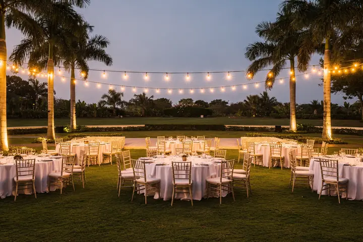 A resort lawn set for an evening reception, round tables with white linens under string lights and palms at dusk