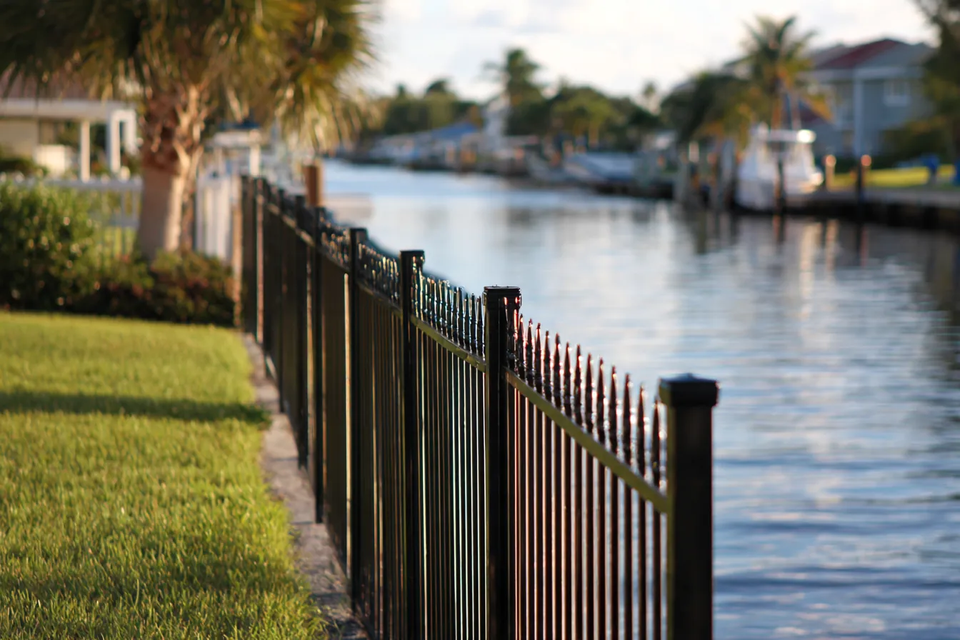 Black aluminum open picket fence along the back of a lawn above a concrete seawall, calm canal water beyond
