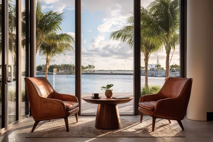 Two leather chairs and a round walnut table before tall windows onto a calm harbor and palms