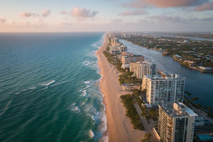 Golden-hour aerial view along an Atlantic coast South Florida city, a wide beach, palms and high-rise condos beside a waterway