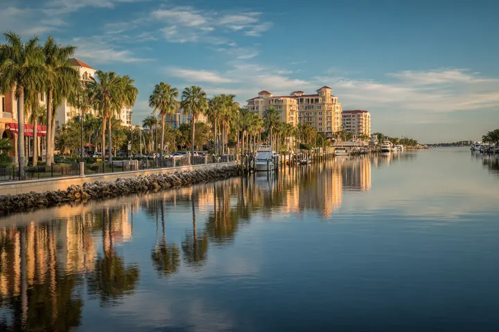 Golden-hour view across a wide calm river to a low-rise Southwest Florida downtown lined with royal palms