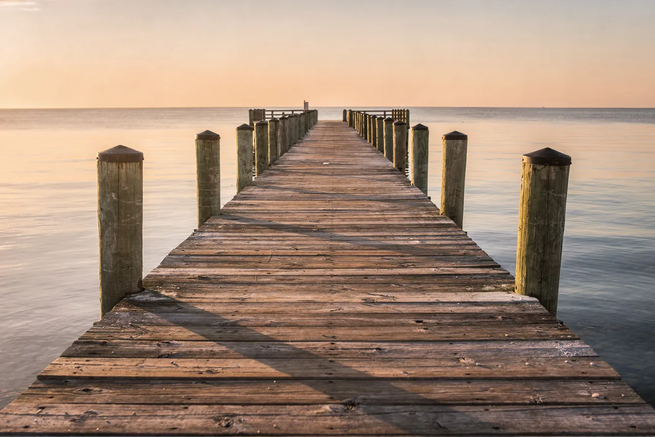 A long wooden fishing pier stretching into a calm Gulf at golden hour, no one on it