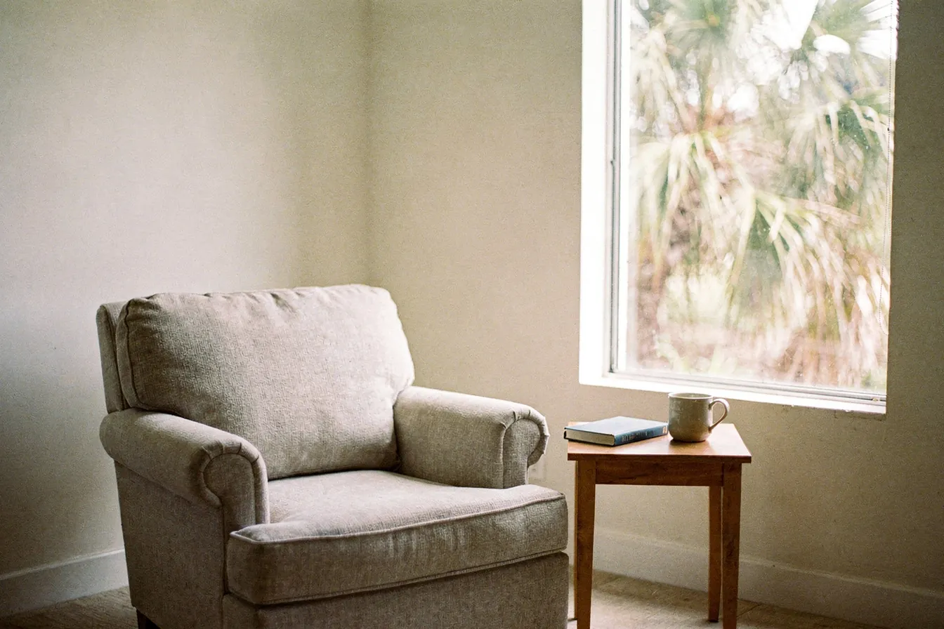A reading chair beside a tall window with palms outside in soft afternoon light