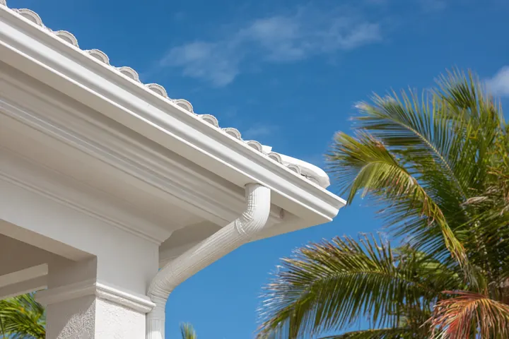 A white seamless gutter and downspout at the corner of a tile roof, palms against a blue sky