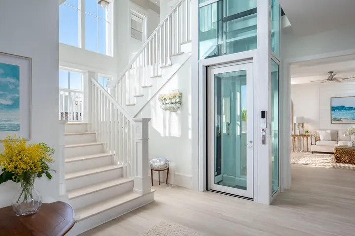 A glass-door residential elevator beside a white staircase in a bright two-story foyer