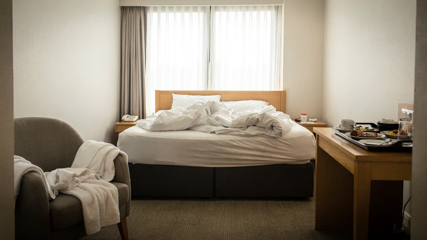A hotel guest room seen straight-on from the doorway, the bed unmade, towels and a room-service tray left on the chair and desk
