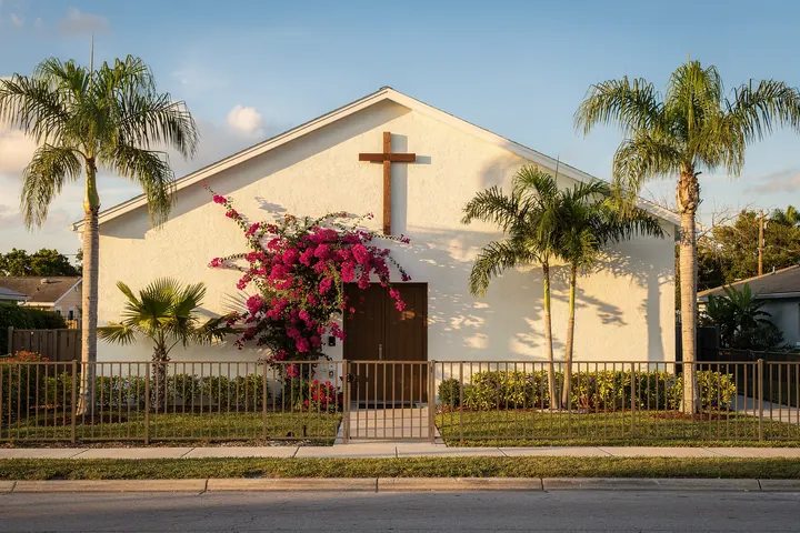 Una pequeña iglesia blanca con una cruz de madera, buganvilla en flor y palmeras detrás de una reja baja, con luz de la tarde