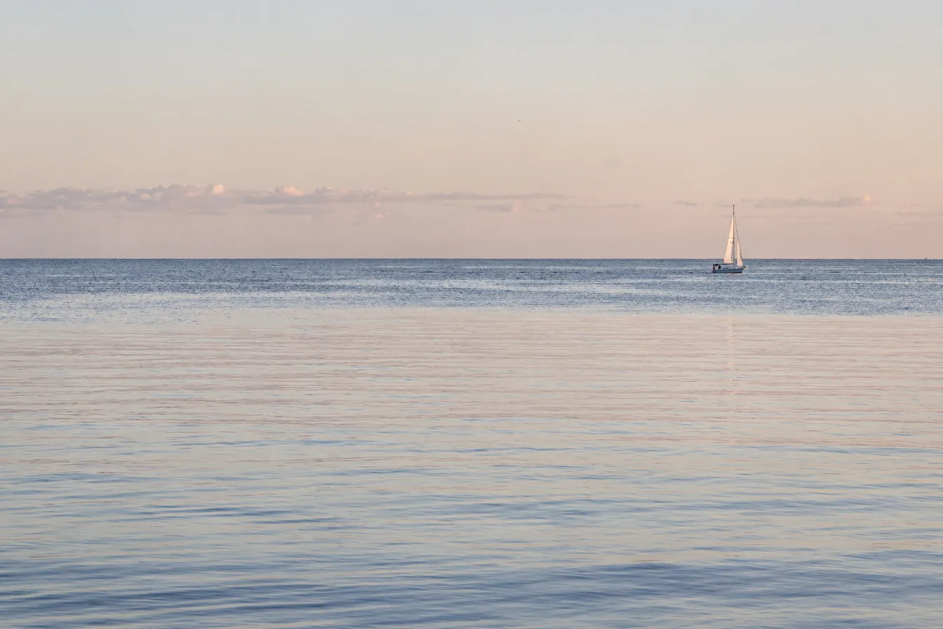 An open ocean horizon at dawn with a single sailboat far out on calm water