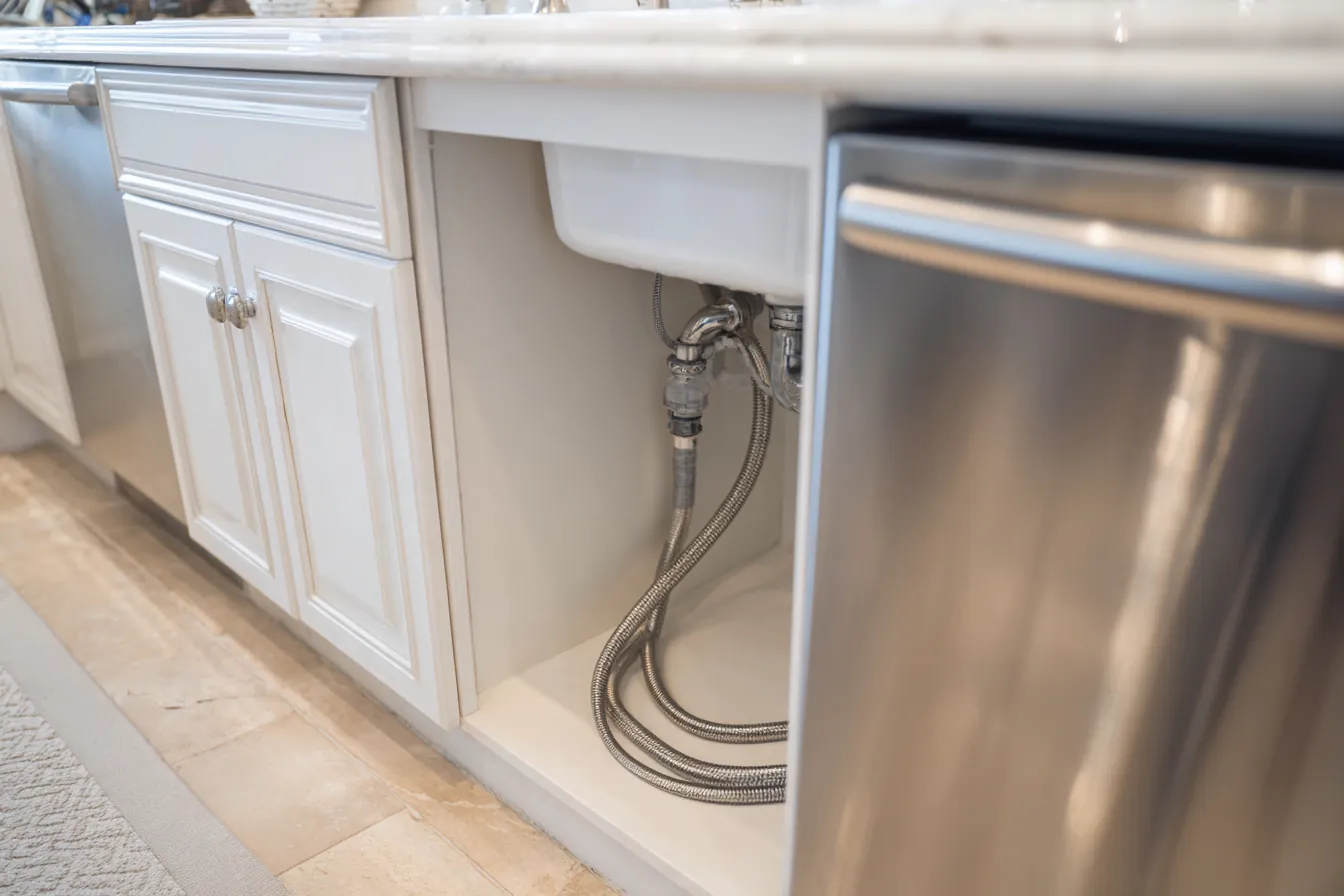 Open sink base cabinet beside a newly installed dishwasher, showing a braided water supply line and a chrome shutoff valve