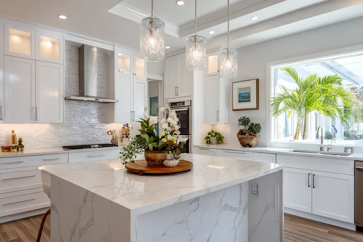 A bright white kitchen with a marble waterfall island, pendant lights and a window onto palms