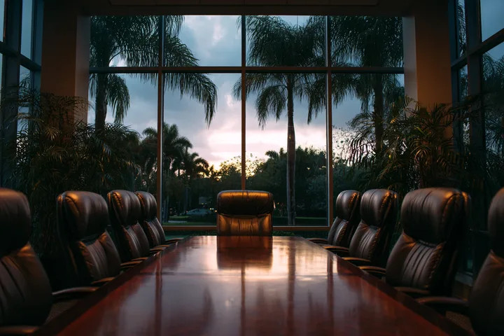 A law office conference room with leather chairs around a long polished table and tall windows onto palms at dusk