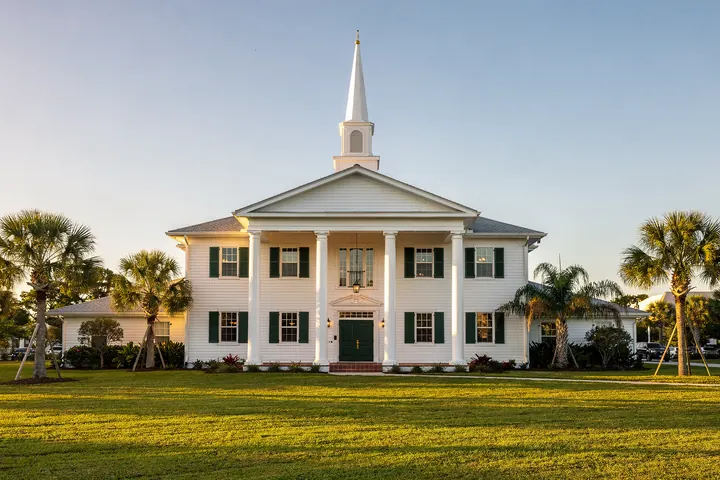 A white colonial style church with a tall steeple, a columned portico and green shutters on a broad lawn with palms in late afternoon light
