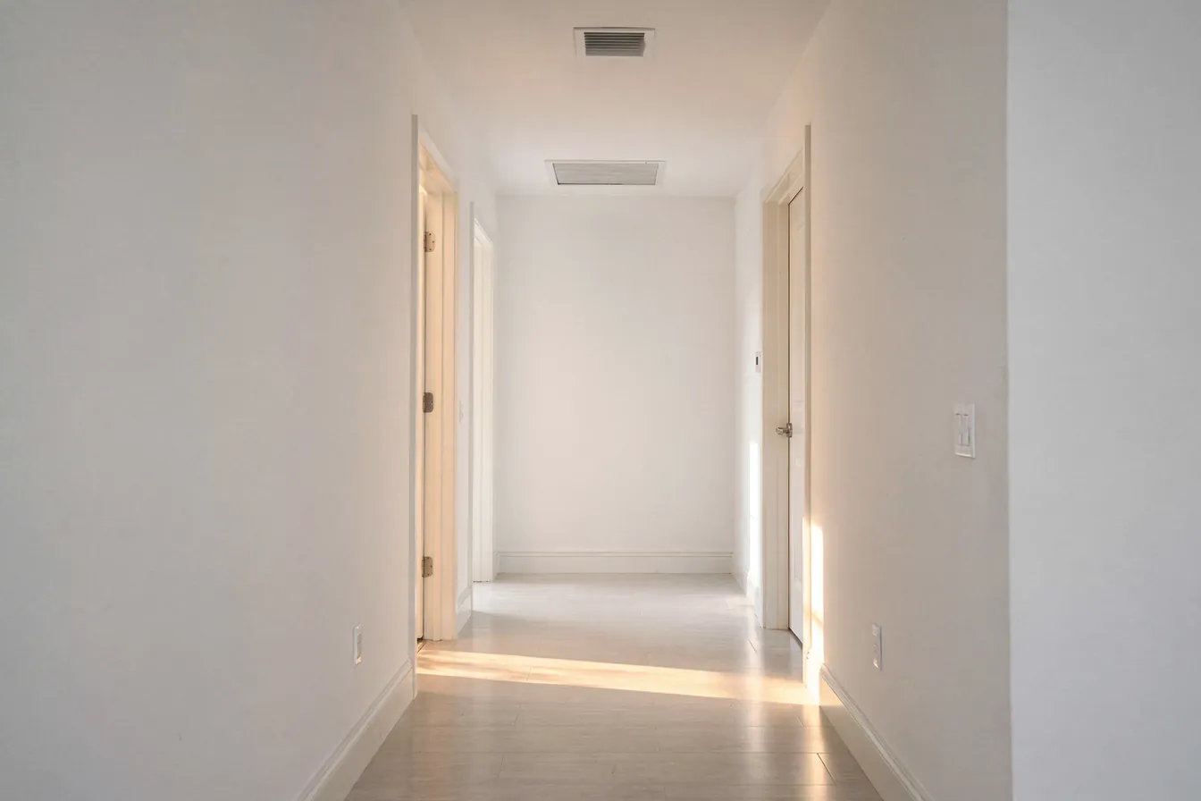 A quiet home hallway with a ceiling vent and soft afternoon light on white walls
