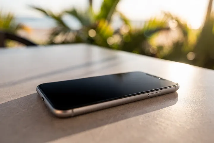 A smartphone with a dark screen resting on a sunlit outdoor cafe table, palms soft in the background