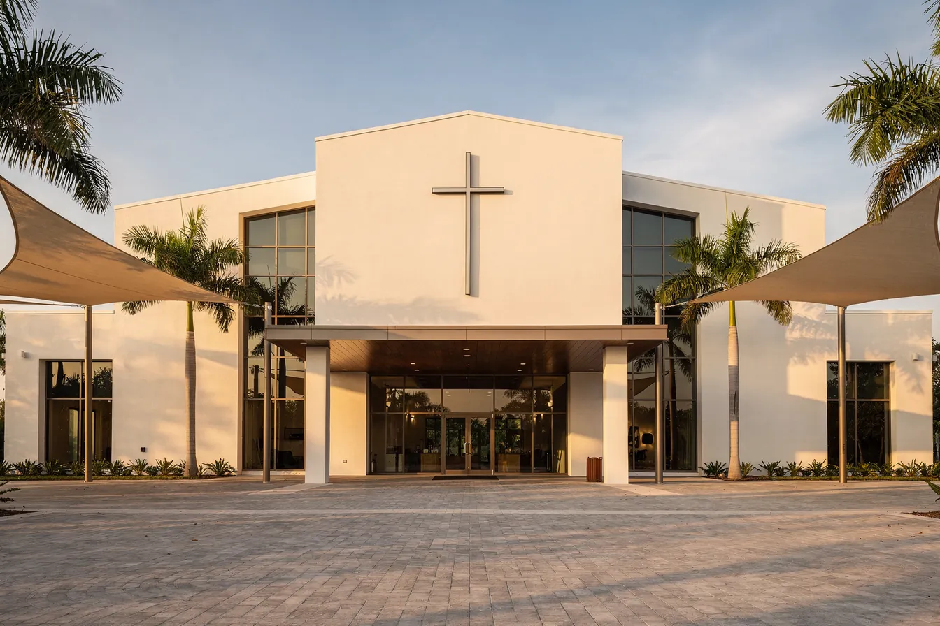 A modern white church with a plain cross on its facade, a glass entry under a wide canopy, palms and shade sails in late afternoon light