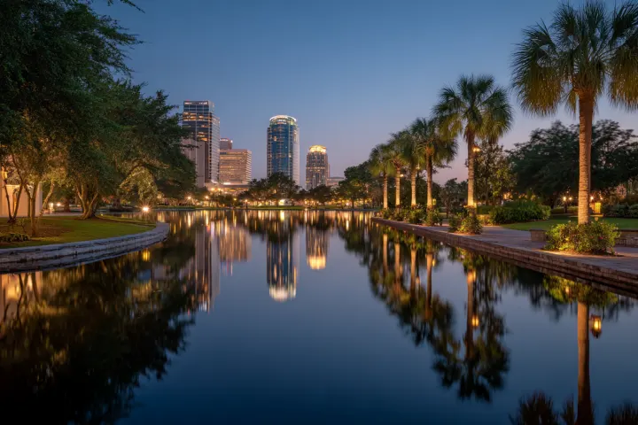A calm city lake at dusk ringed by palms and oaks, generic downtown towers lit beyond the water
