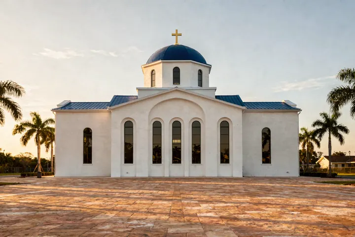 A white Byzantine style church with a blue dome and a gold cross, arched windows and palms in late afternoon light