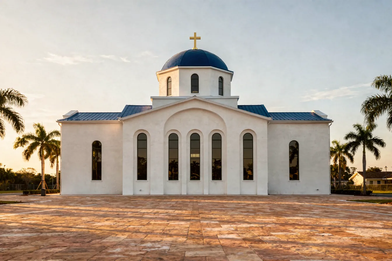 A white Byzantine style church with a blue dome and a gold cross, arched windows and palms in late afternoon light