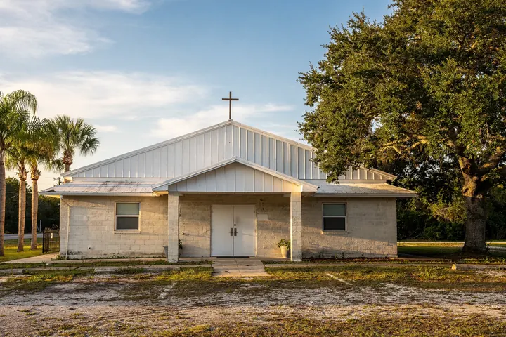 A modest concrete block church with a white metal gable, a plain cross and double doors under a large live oak in late afternoon light