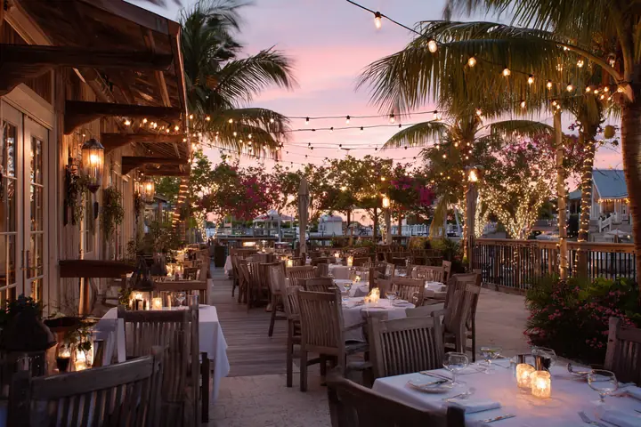 A softly lit restaurant terrace at dusk, set tables with candles under string lights and palms