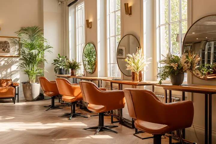 A bright hair salon in soft morning light, a row of styling chairs and round mirrors, plants by tall windows