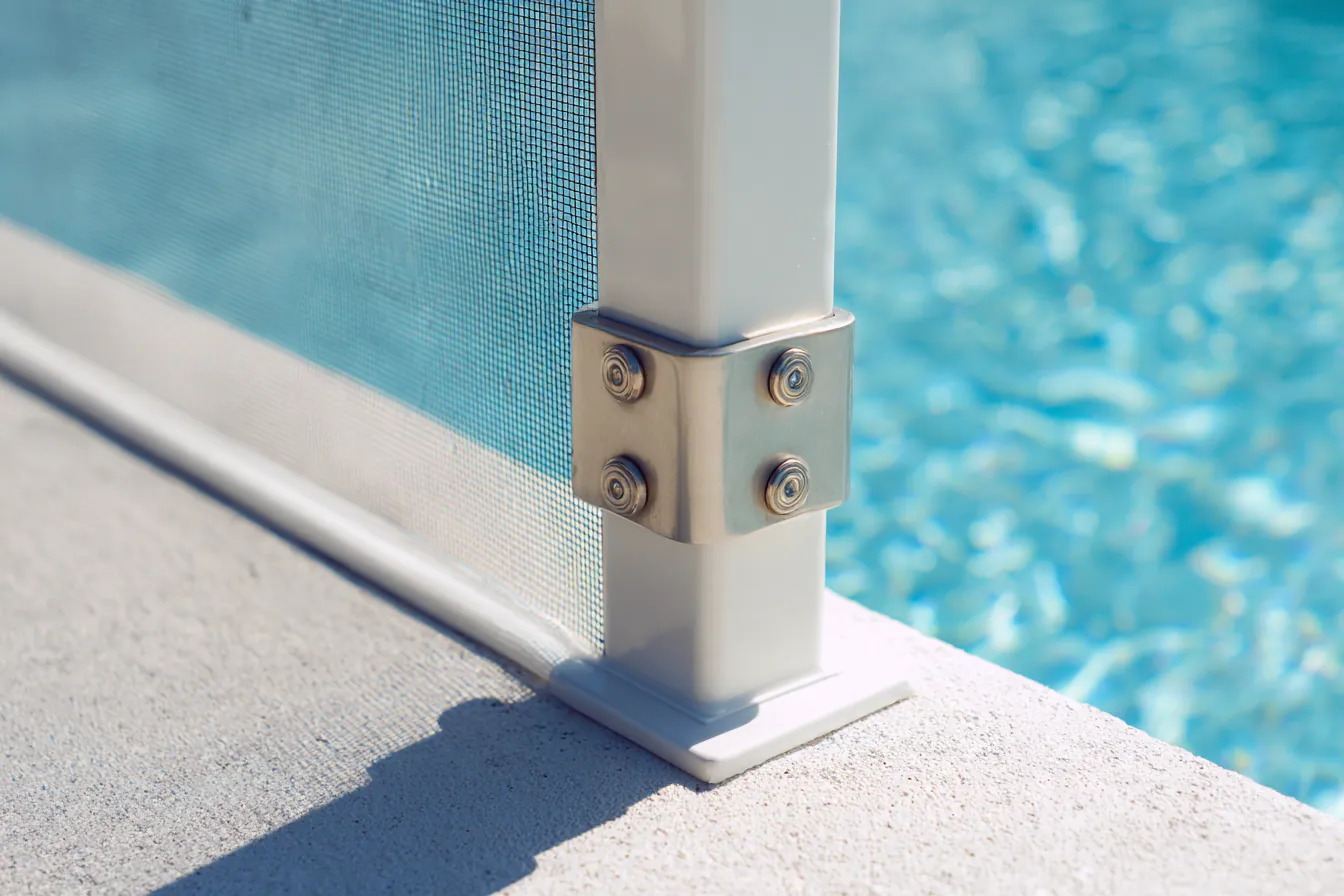 Close view of a white extruded aluminum pool cage corner post bolted to a pale concrete pool deck, gray screen mesh stretched taut behind it