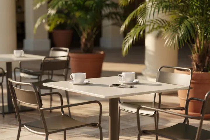 A sunlit cafe terrace with empty tables and potted palms, a phone lying beside two coffee cups