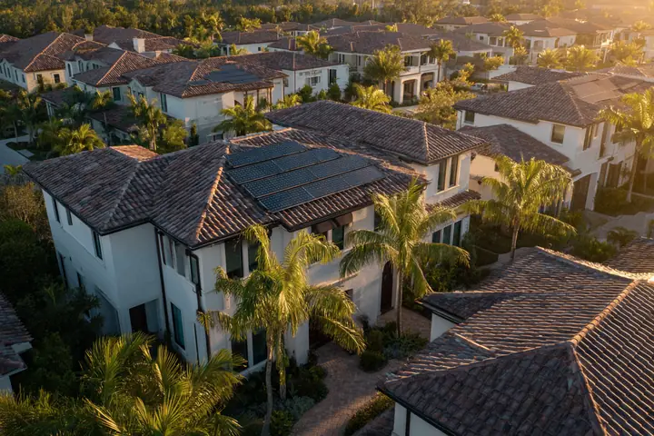 An aerial view over Southwest Florida tile rooftops at golden hour, several carrying dark solar arrays, palms between the homes