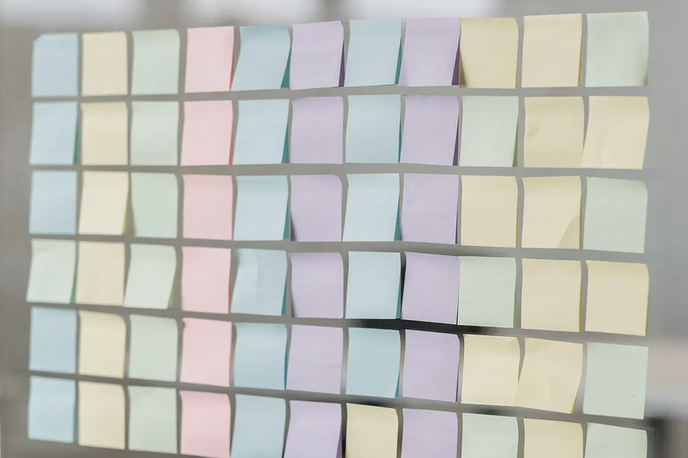 A glass wall of blank pastel sticky notes in neat columns, markers on the ledge below