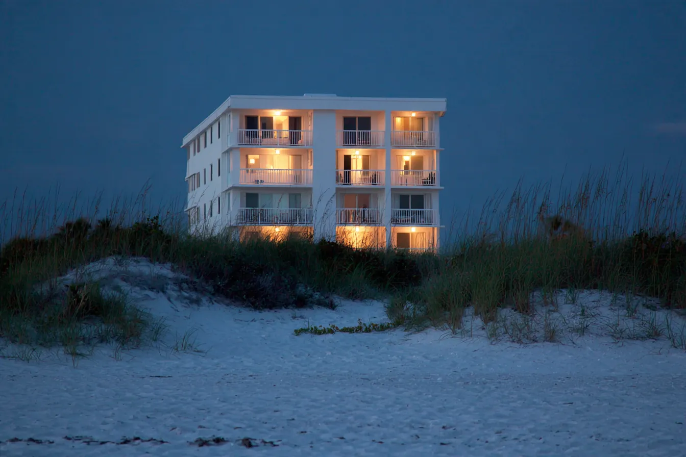 A low-rise beachfront condominium at dusk seen across a sand dune with sea oats, its windows dim and softly tinted