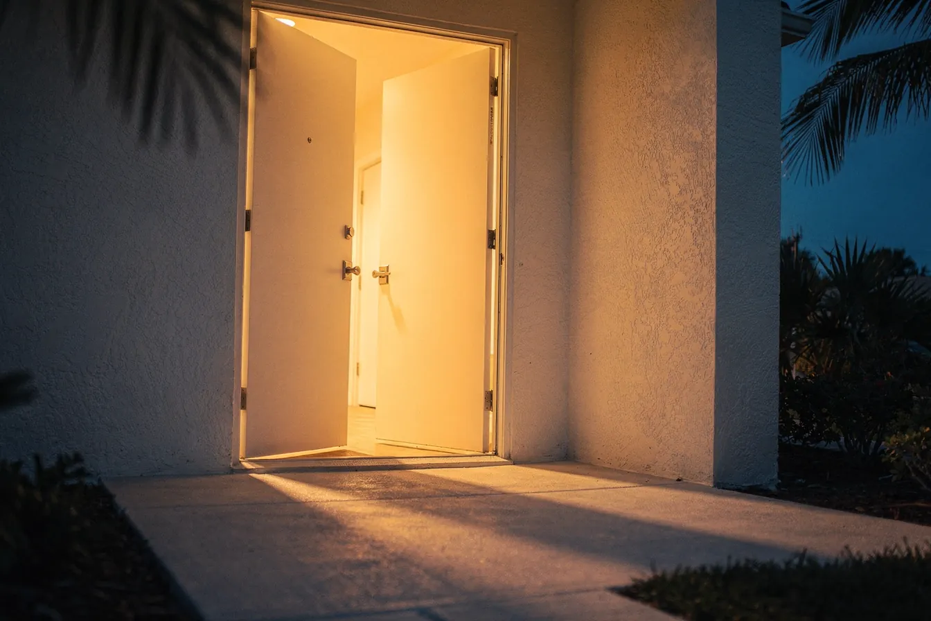 A wide open doorway at blue hour, warm light spilling across a pale stucco path