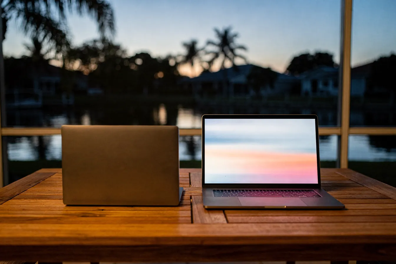 A laptop open on a teak table on a screened lanai at dusk, a calm canal and palms beyond
