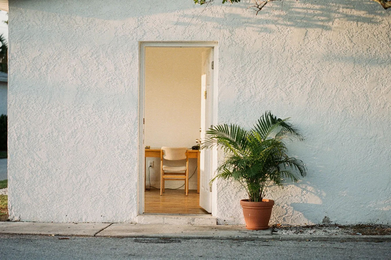 The open door of a small white stucco office on a quiet Southwest Florida street, a desk inside and a potted palm by the step