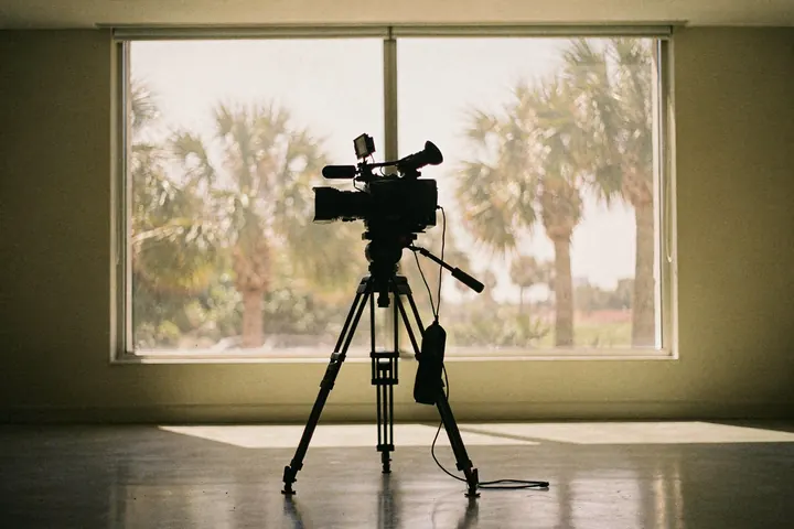 A plain video camera on a tripod facing an empty sunlit room, palms beyond the window