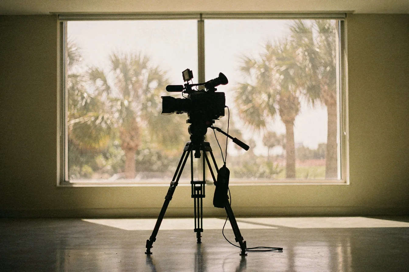A plain video camera on a tripod facing an empty sunlit room, palms beyond the window