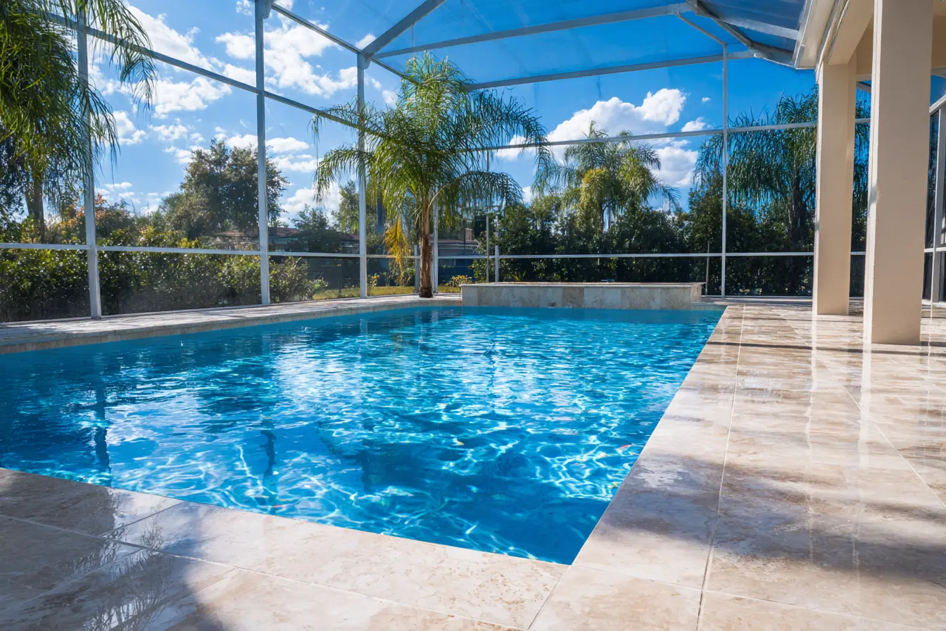 A freshly cleaned travertine pool deck under a screen enclosure, the pool water bright blue