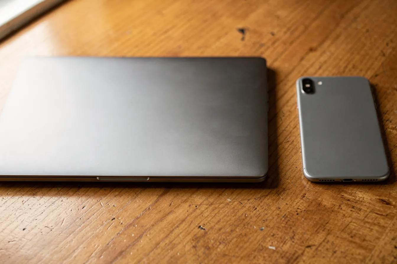 A smartphone and a laptop side by side on a wooden desk, both screens dark